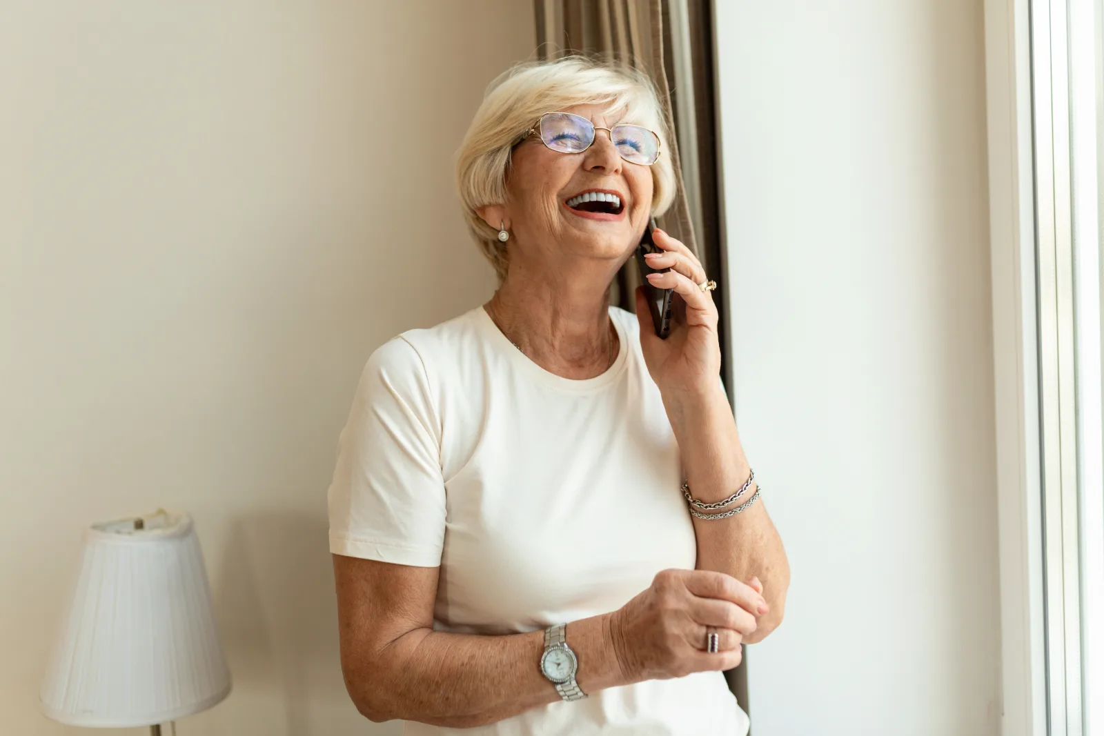 Seniorin telefoniert lächelnd zu Hause am Fenster